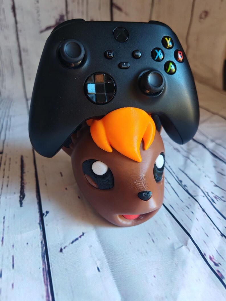Pode incluir: Um controlador Xbox preto est&aacute; em cima da cabe&ccedil;a de um personagem Pokemon impresso em 3D de cor marrom e laranja.