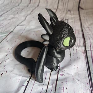 K&ouml;nnte beinhalten: Eine schwarze, 3D-gedruckte Drachenfigur mit gro&szlig;en gr&uuml;nen Augen. Der Drache hat einen runden K&ouml;rper, gro&szlig;e Ohren und einen gebogenen Schwanz. Die Fl&uuml;gel sind ausgebreitet. Die Figur steht vor einem wei&szlig;-grau gestreiften Hintergrund.