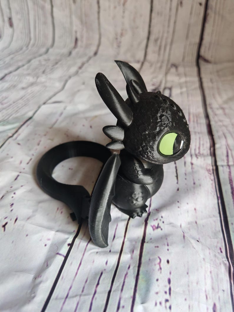 K&ouml;nnte beinhalten: Eine schwarze, 3D-gedruckte Drachenfigur mit gro&szlig;en gr&uuml;nen Augen. Der Drache hat einen runden K&ouml;rper, gro&szlig;e Ohren und einen gebogenen Schwanz. Die Fl&uuml;gel sind ausgebreitet. Die Figur steht vor einem wei&szlig;-grau gestreiften Hintergrund.