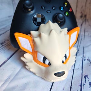 Peut inclure: Un support d'impression 3D pour une manette de jeu vidéo, en forme de renard blanc et orange avec des yeux noirs. Le support est conçu pour tenir une manette Xbox noire.