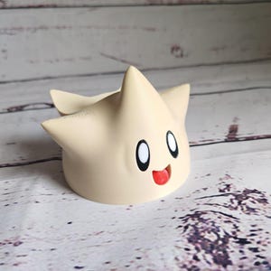 May include: A beige 3D printed figurine of a star-shaped character with a smiling face and large eyes. The character has a red tongue sticking out.