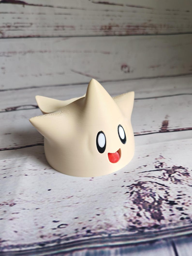 May include: A beige 3D printed figurine of a star-shaped character with a smiling face and large eyes. The character has a red tongue sticking out.
