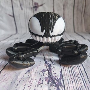 May include: Black and white 3D printed toy of Venom, a fictional character from Marvel Comics. The toy has a spider-like body with multiple legs and a head with a white face and black eyes.