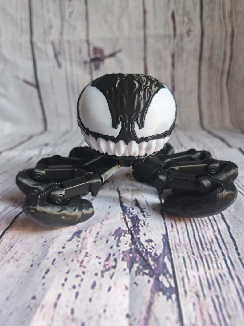 May include: Black and white 3D printed toy of Venom, a fictional character from Marvel Comics. The toy has a spider-like body with multiple legs and a head with a white face and black eyes.