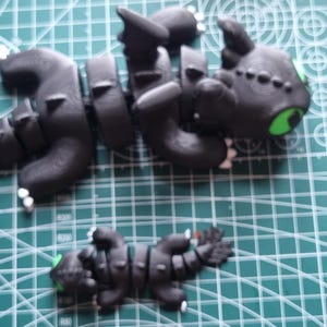 May include: Two black and green dragon figurines. The larger dragon is in a flying pose with its wings spread out. The smaller dragon is lying on its belly. Both dragons have green eyes.