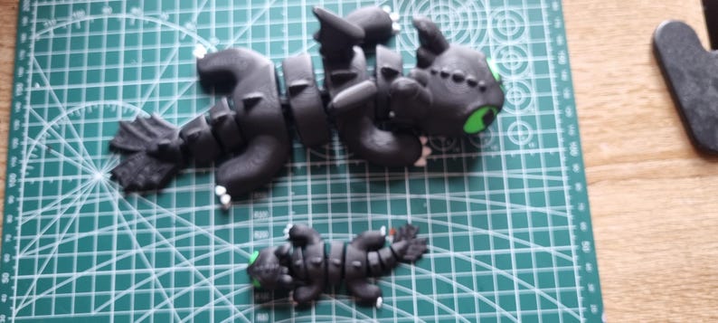 May include: Two black and green dragon figurines. The larger dragon is in a flying pose with its wings spread out. The smaller dragon is lying on its belly. Both dragons have green eyes.
