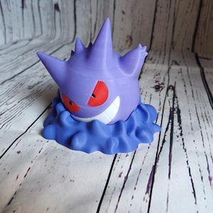 Może przedstawiać: Fioletowa i biała figurka 3D postaci Pokemon, Gengar, z czerwonymi oczami i przebiegłym uśmiechem. Figurka stoi na chmurowatej podstawie.