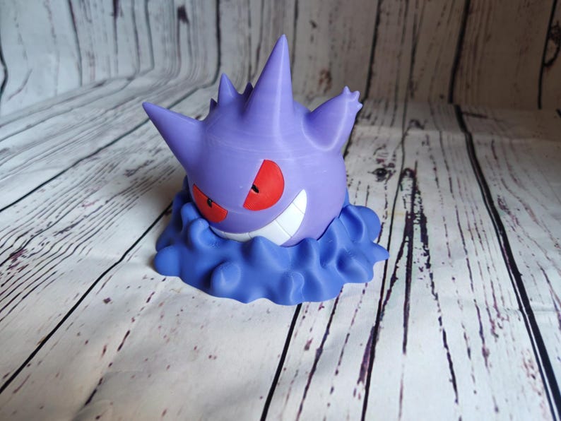 Może przedstawiać: Fioletowa i biała figurka 3D postaci Pokemon, Gengar, z czerwonymi oczami i przebiegłym uśmiechem. Figurka stoi na chmurowatej podstawie.
