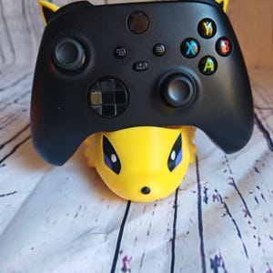 Peut inclure: Une manette Xbox noire avec des boutons jaunes et noirs est pos&eacute;e sur un support de personnage Pikachu jaune.