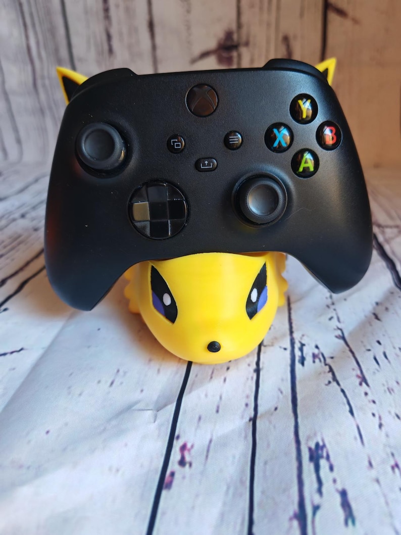Peut inclure: Une manette Xbox noire avec des boutons jaunes et noirs est pos&eacute;e sur un support de personnage Pikachu jaune.