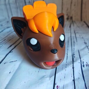 Pode incluir: Uma figura de Pokemon impressa em 3D de cor marrom e laranja com uma grande juba laranja e uma l&iacute;ngua vermelha que se projeta.