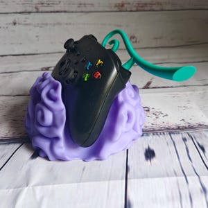 Op de afbeelding: Een paarse 3D-geprinte standaard in de vorm van een wolk met een zwarte videogamecontroller erop. Een turquoise plastic headset-houder is aan de standaard bevestigd.