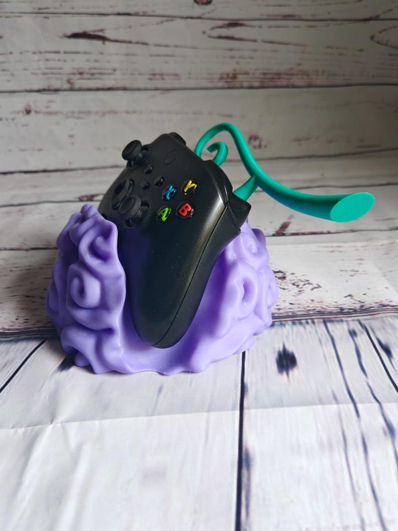 Op de afbeelding: Een paarse 3D-geprinte standaard in de vorm van een wolk met een zwarte videogamecontroller erop. Een turquoise plastic headset-houder is aan de standaard bevestigd.