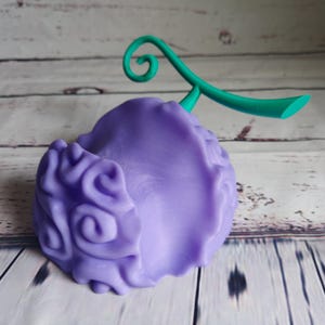 Op de afbeelding: Een 3D-geprint figuurtje van de Gum-Gum-vrucht uit de anime One Piece, in de kleuren paars en groen. De vrucht heeft een swirl-patroon en een groene steel.