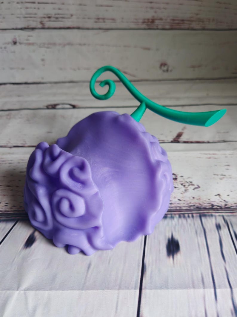 Op de afbeelding: Een 3D-geprint figuurtje van de Gum-Gum-vrucht uit de anime One Piece, in de kleuren paars en groen. De vrucht heeft een swirl-patroon en een groene steel.