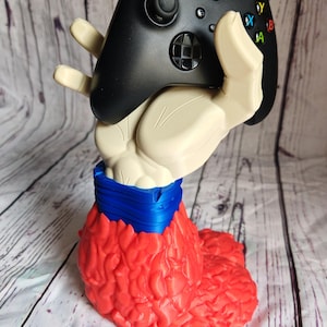 Pode incluir: Um controle Xbox preto repousa sobre um suporte em forma de m&atilde;o bege. O suporte tem uma faixa azul ao redor do pulso e uma base vermelha texturizada. O controle possui bot&otilde;es coloridos e joysticks.