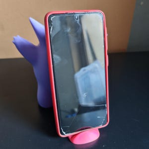 Może przedstawiać: Czarny smartfon w czerwonym etui spoczywa na małym, r&oacute;żowym stojaku. Fioletowa, stylizowana figurka podtrzymuje telefon od tyłu. Ekran telefonu jest pęknięty.