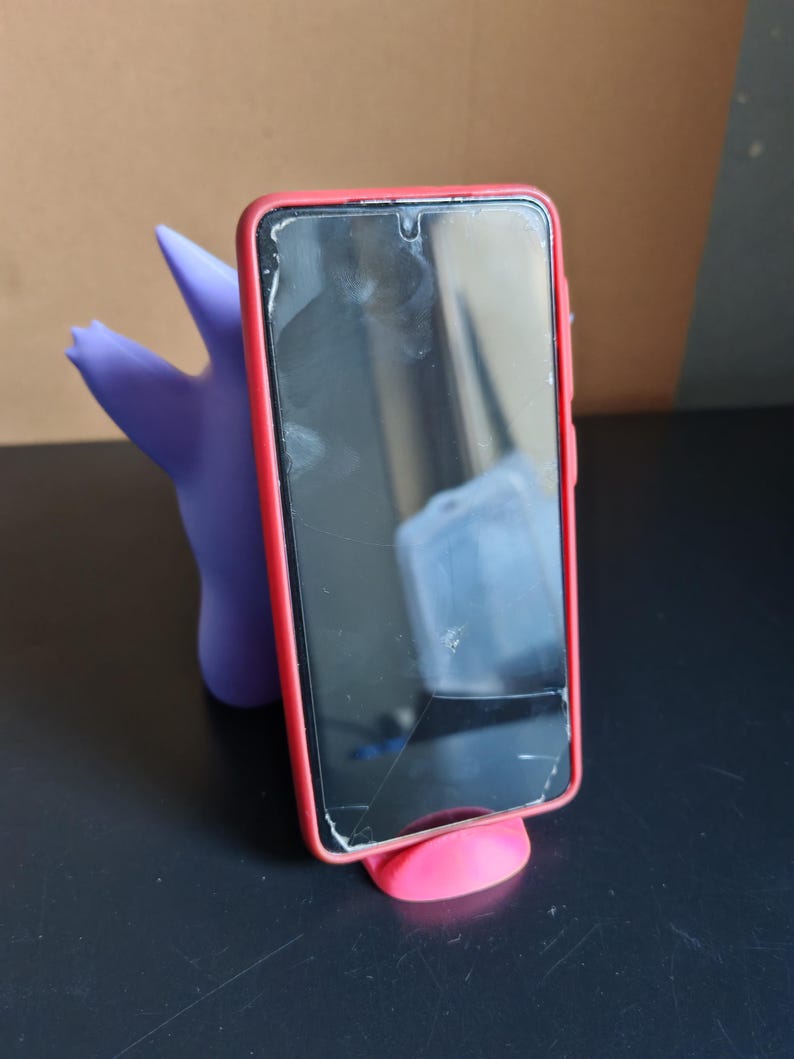 Może przedstawiać: Czarny smartfon w czerwonym etui spoczywa na małym, r&oacute;żowym stojaku. Fioletowa, stylizowana figurka podtrzymuje telefon od tyłu. Ekran telefonu jest pęknięty.