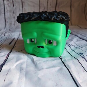 K&ouml;nnte beinhalten: Ein gr&uuml;ner Blumentopf im Cartoon-Stil in Form eines Gesichts mit schwarzen Haaren. Der Topf hat wei&szlig;e Augen, eine kleine schwarze Nase und einen leicht traurigen Ausdruck. Der Topf steht auf einer wei&szlig;-grauen Oberfl&auml;che.