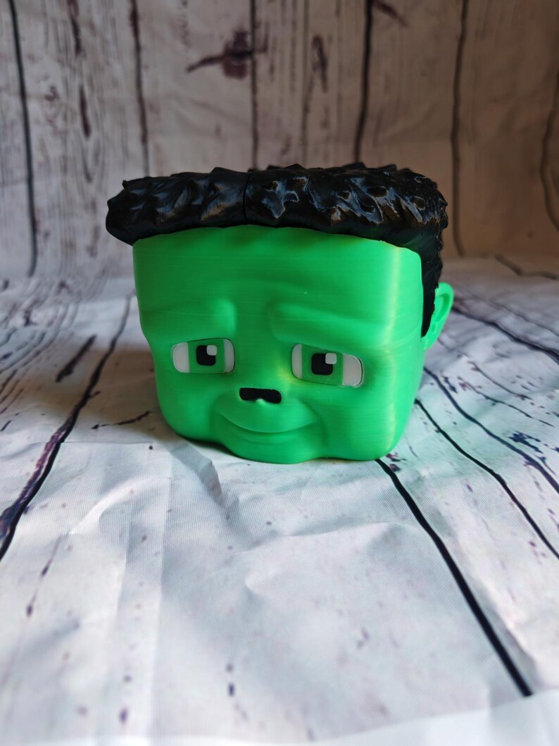 K&ouml;nnte beinhalten: Ein gr&uuml;ner Blumentopf im Cartoon-Stil in Form eines Gesichts mit schwarzen Haaren. Der Topf hat wei&szlig;e Augen, eine kleine schwarze Nase und einen leicht traurigen Ausdruck. Der Topf steht auf einer wei&szlig;-grauen Oberfl&auml;che.