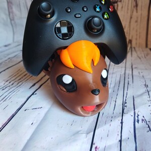 Pode incluir: Um controlador Xbox preto est&aacute; em cima da cabe&ccedil;a de um personagem impresso em 3D marrom e laranja. O personagem tem olhos grandes e uma l&iacute;ngua vermelha para fora.