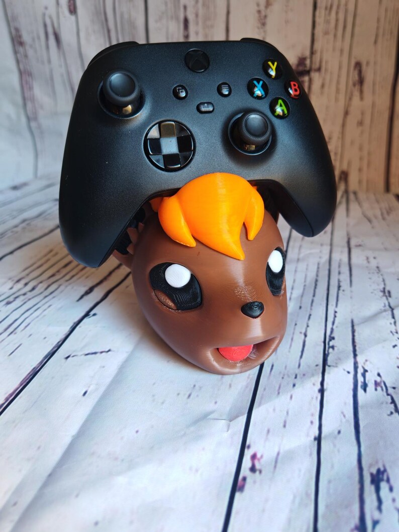 Pode incluir: Um controlador Xbox preto est&aacute; em cima da cabe&ccedil;a de um personagem impresso em 3D marrom e laranja. O personagem tem olhos grandes e uma l&iacute;ngua vermelha para fora.