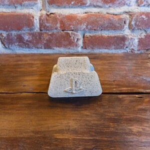May include: A white concrete PlayStation controller stand with a PlayStation logo. The stand is on a wooden surface.