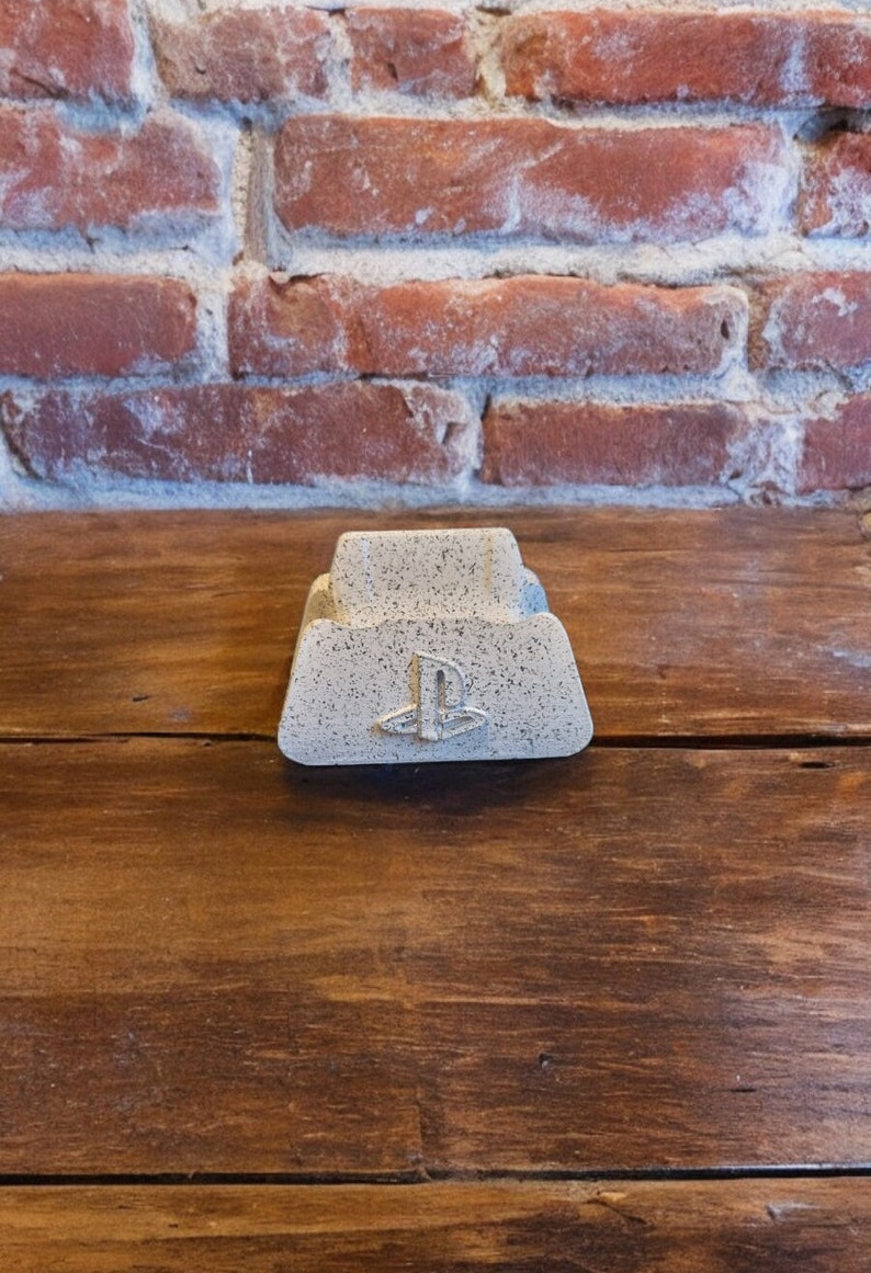 May include: A white concrete PlayStation controller stand with a PlayStation logo. The stand is on a wooden surface.
