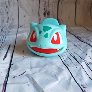 Puede incluir: Una figurita de pl&aacute;stico azul claro y verde de un personaje de Pokemon con ojos rojos. La figurita tiene forma de un peque&ntilde;o dinosaurio con una boca ancha y una cola peque&ntilde;a.