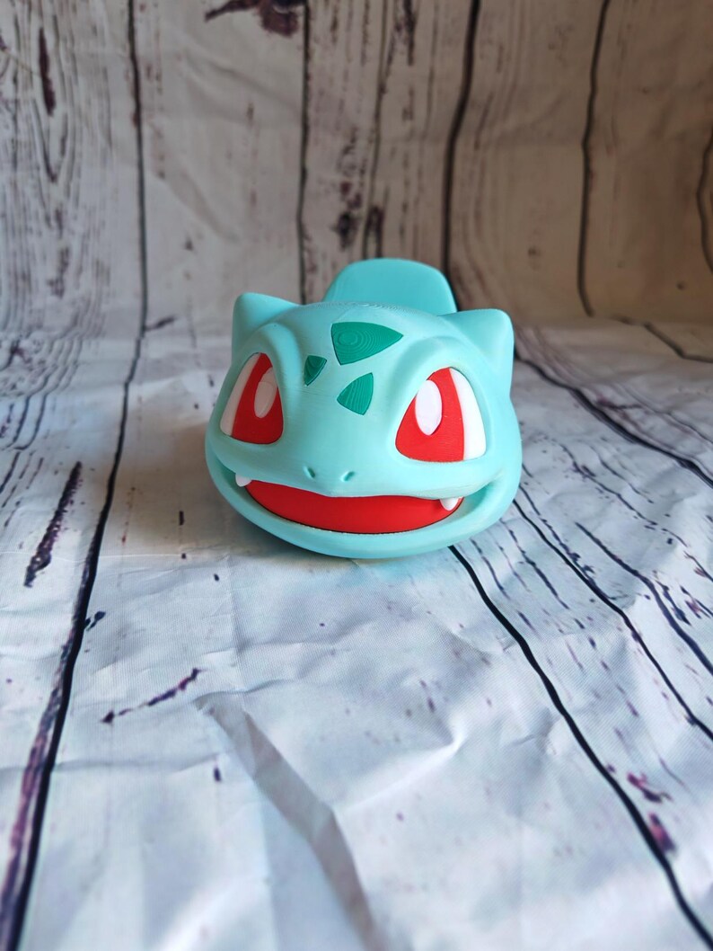 Puede incluir: Una figurita de pl&aacute;stico azul claro y verde de un personaje de Pokemon con ojos rojos. La figurita tiene forma de un peque&ntilde;o dinosaurio con una boca ancha y una cola peque&ntilde;a.
