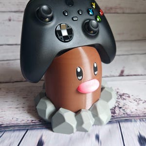 Op de afbeelding: Een bruin en grijs 3D-geprint Pok&eacute;mon-personage, dat lijkt op een grondtype, houdt een zwarte Xbox-controller vast.