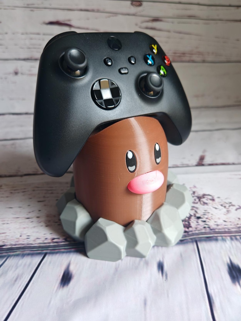 Op de afbeelding: Een bruin en grijs 3D-geprint Pok&eacute;mon-personage, dat lijkt op een grondtype, houdt een zwarte Xbox-controller vast.