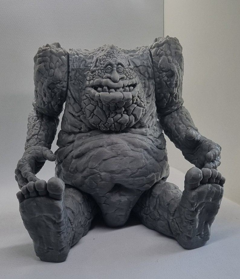 May include: A gray, stone-like figure with a large, round belly and a face with a wide mouth and small eyes. The figure is sitting with its legs crossed and its arms resting on its knees.