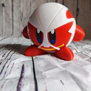Pode incluir: Uma figura vermelha e branca de um personagem com grandes olhos azuis, pequenos bra&ccedil;os e p&eacute;s vermelhos. O personagem tem uma estrutura branca em forma de capacete com detalhes vermelhos. A figura est&aacute; sobre uma superf&iacute;cie estampada em branco e cinza.