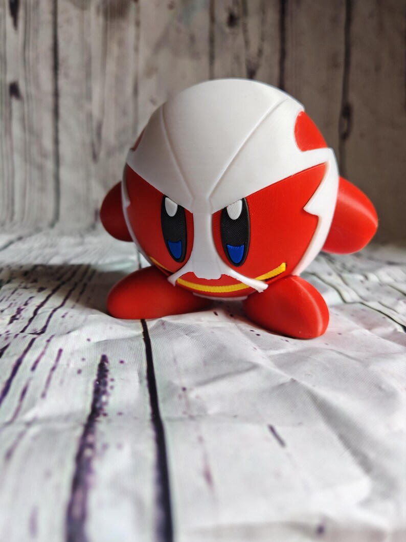 Pode incluir: Uma figura vermelha e branca de um personagem com grandes olhos azuis, pequenos bra&ccedil;os e p&eacute;s vermelhos. O personagem tem uma estrutura branca em forma de capacete com detalhes vermelhos. A figura est&aacute; sobre uma superf&iacute;cie estampada em branco e cinza.