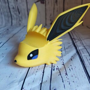 Peut inclure: Une figurine Eevee imprim&eacute;e en 3D de couleur jaune et noire avec un &oelig;il bleu. La figurine est pos&eacute;e la t&ecirc;te inclin&eacute;e sur le c&ocirc;t&eacute; et les oreilles dress&eacute;es.