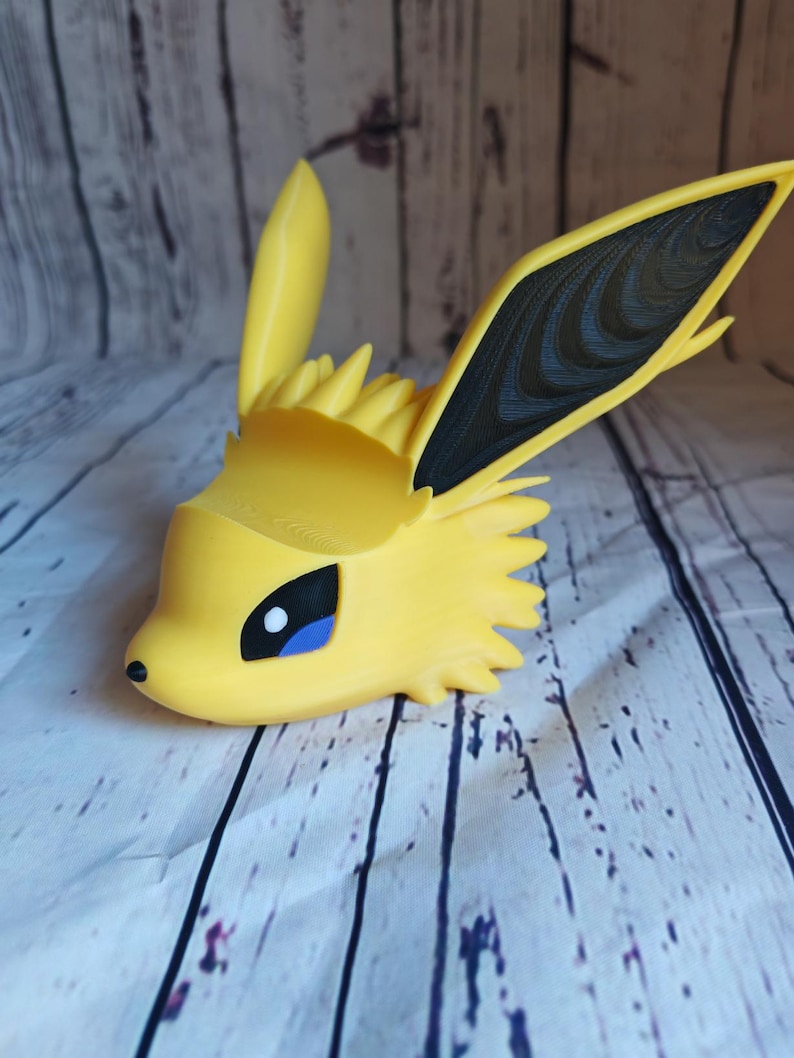 Peut inclure: Une figurine Eevee imprim&eacute;e en 3D de couleur jaune et noire avec un &oelig;il bleu. La figurine est pos&eacute;e la t&ecirc;te inclin&eacute;e sur le c&ocirc;t&eacute; et les oreilles dress&eacute;es.