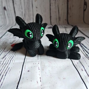 May include: Two black crocheted plush toys shaped like dragons with green eyes. The dragons are sitting on a white wooden surface.