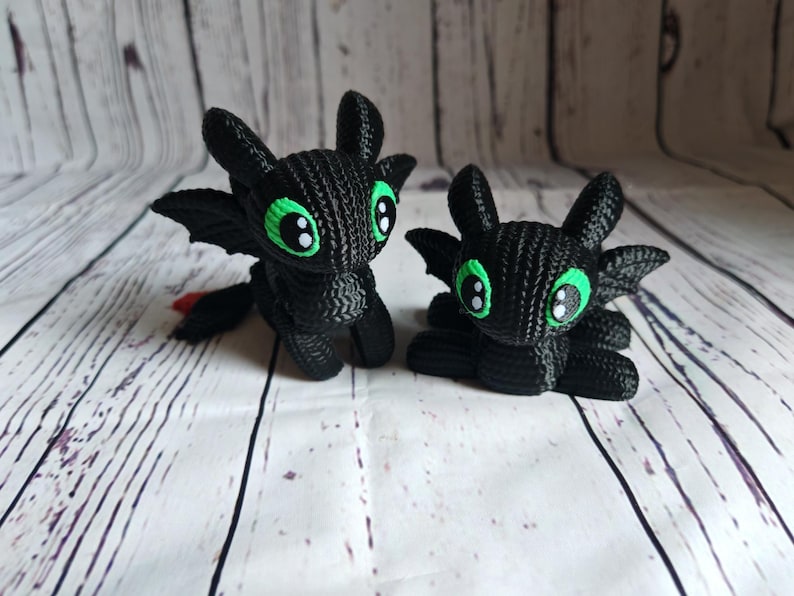 May include: Two black crocheted plush toys shaped like dragons with green eyes. The dragons are sitting on a white wooden surface.