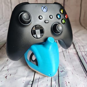 Op de afbeelding: Zwarte Xbox-controller op een blauw en tan Lapras-thema controllerstandaard. De standaard is 3D-geprint en ontworpen om de controller vast te houden. De controller heeft gekleurde knoppen en twee joysticks.