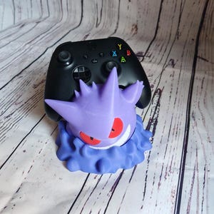 Może przedstawiać: Fioletowa i niebieska postać Pok&eacute;mona Gengar trzymająca czarny kontroler Xbox. Kontroler ma kolorowe przyciski oznaczone literami 'A', 'B', 'X' i 'Y'.