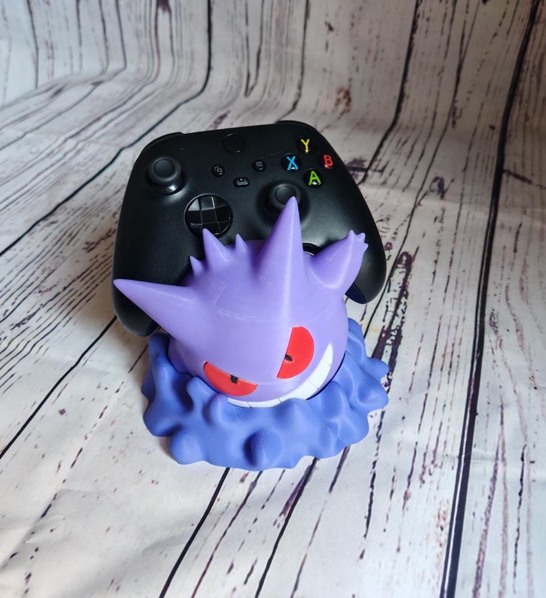Może przedstawiać: Fioletowa i niebieska postać Pok&eacute;mona Gengar trzymająca czarny kontroler Xbox. Kontroler ma kolorowe przyciski oznaczone literami 'A', 'B', 'X' i 'Y'.