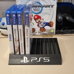 Può includere: Un supporto per videogiochi in plastica nera con logo PS5. Il supporto ha slot per contenere custodie di videogiochi. Quattro custodie di videogiochi sono esposte nel supporto. Le custodie sono per i giochi PS4: Killzone Shadow Fall, Dragon Ball Xenoverse, Ghost of Tsushima e The Sims 4.