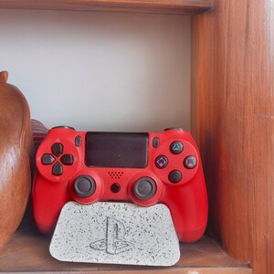 May include: A red PlayStation 4 controller is sitting on a white and gray PlayStation themed stand. The stand has the PlayStation logo on it.