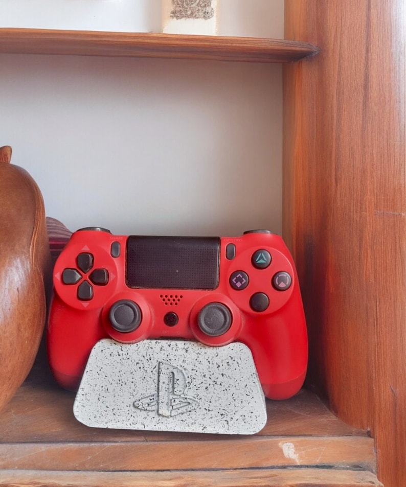 May include: A red PlayStation 4 controller is sitting on a white and gray PlayStation themed stand. The stand has the PlayStation logo on it.