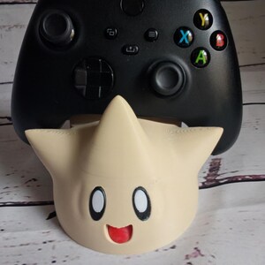 May include: A black Xbox controller with a yellow, blue, green, and red button is sitting on a beige star-shaped stand with a smiling face.