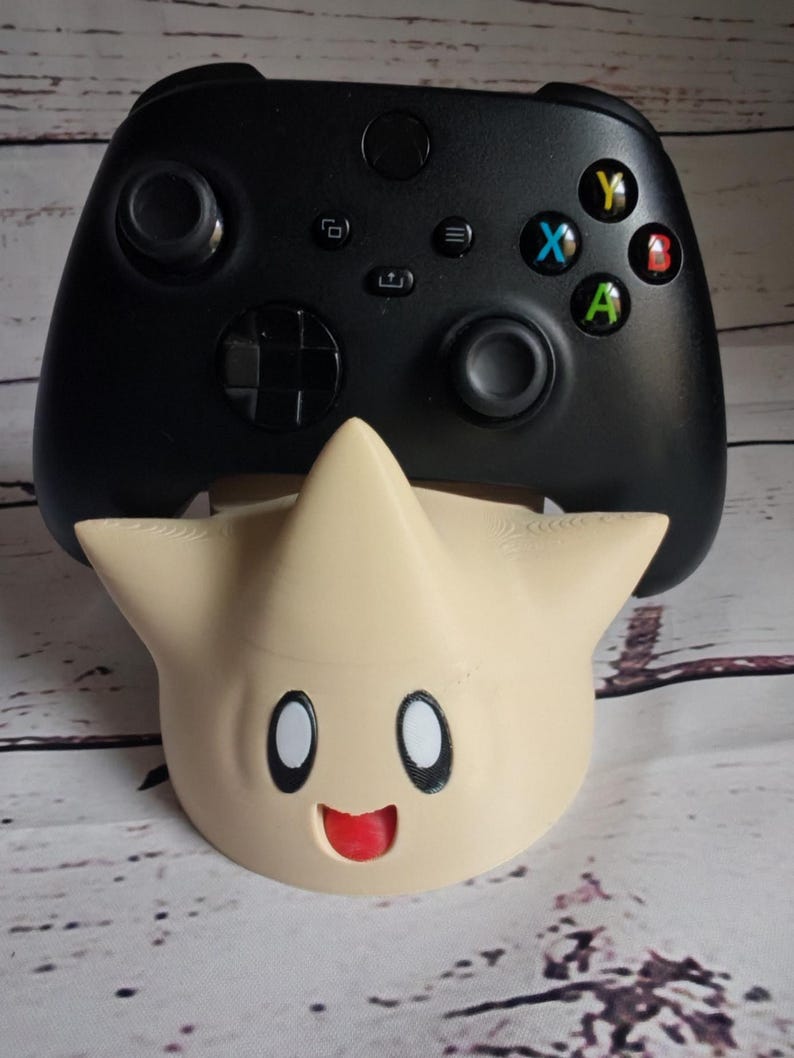 May include: A black Xbox controller with a yellow, blue, green, and red button is sitting on a beige star-shaped stand with a smiling face.