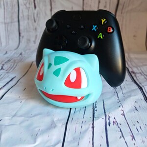 Puede incluir: Un mando Xbox negro con un soporte de Bulbasaur de color azul y verde. El mando tiene botones de colores y una placa frontal negra.