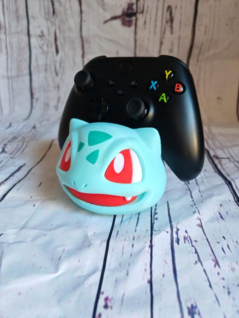 Puede incluir: Un mando Xbox negro con un soporte de Bulbasaur de color azul y verde. El mando tiene botones de colores y una placa frontal negra.