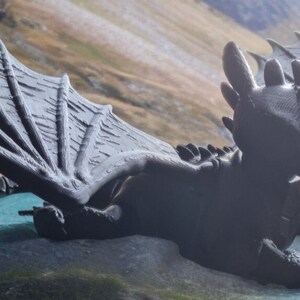 K&ouml;nnte beinhalten: Ein schwarzes 3D-gedrucktes Modell eines Drachen mit ausgebreiteten Fl&uuml;geln. Der Drache liegt auf einer gr&uuml;nen Oberfl&auml;che mit einem Berg im Hintergrund.